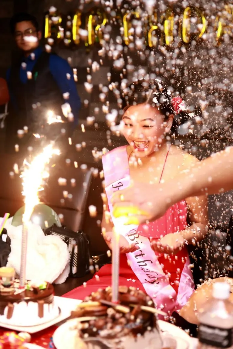 Une jeune femme portant une écharpe d’anniversaire rit sous une pluie de confettis, devant un gâteau à cierges magiques